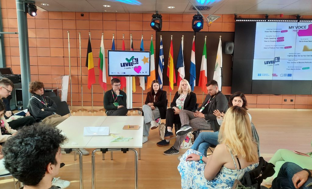 A group of young people sitting around a table. In the background a TV screen shows the LevelUP! 2024 logo.