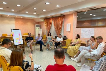 Group of people sitting in a room, discussing. Between them, a whiteboard with post-its.