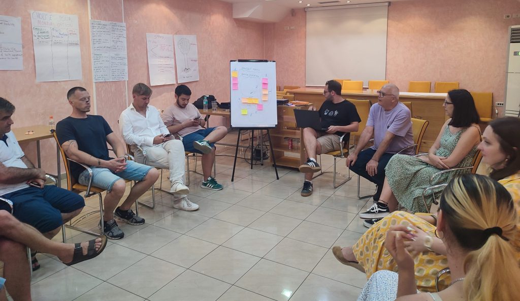 Participants sitting in a room, discussing. Between them, a whiteboard with post-its.