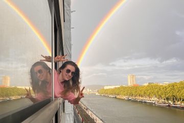 Portait picture of a smiling Kinga peeking outside of a ferry window with two rainbows in the background.