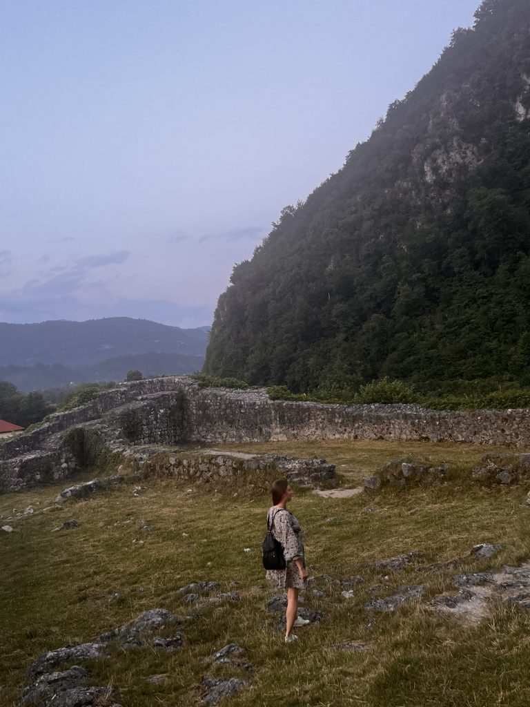 Kinga walks in a rural scenery with ruins and a tall mountain in the background