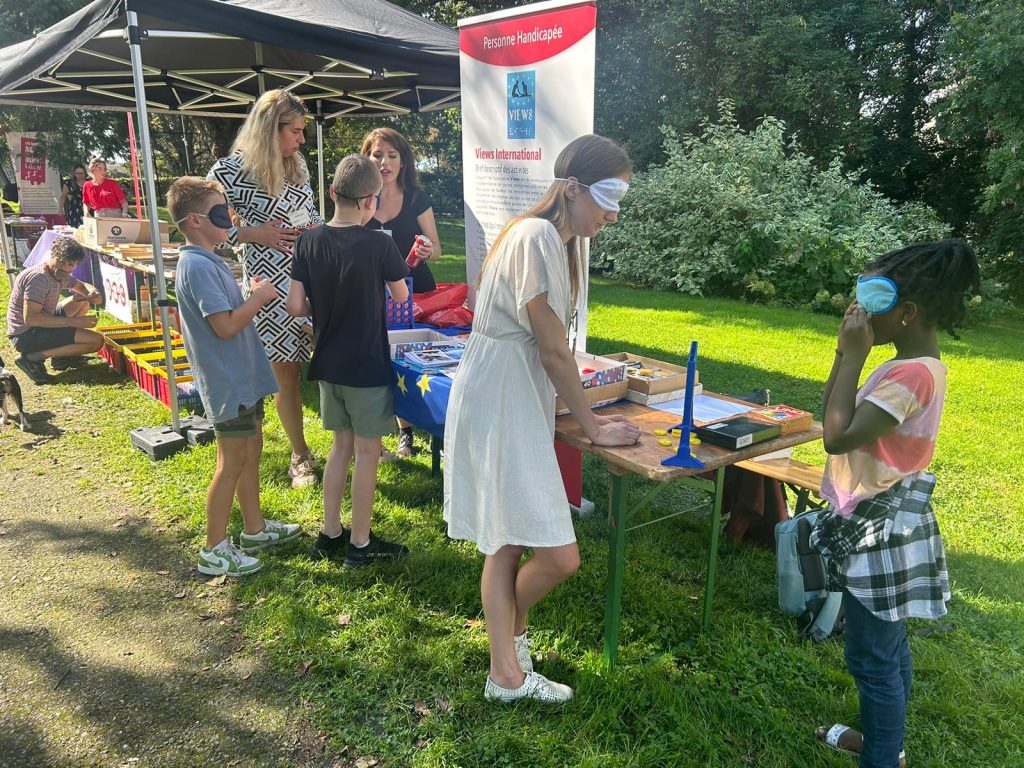 The VIEWS International stand. In the foreground, Kinga plays an adapted table game blindfolded with a child. In the background, Patricia and Sara play with two more blindfolded children.