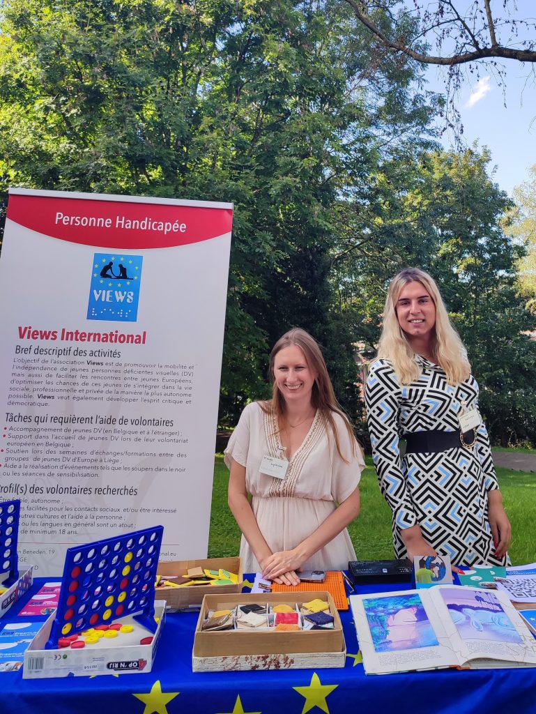 Kinga and Patricia smiling behind the VIEWS International stand, which is covered in adapted board games and books. In the background, a VIEWS International rollup.