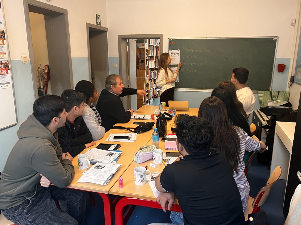 Kinga writing words in French on a blackboard as she gives a lesson. The course's students are listening attentively.