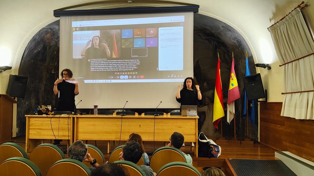 People listening both in the room and online to the organisers as they speak from the podium. A person is translating into sign language